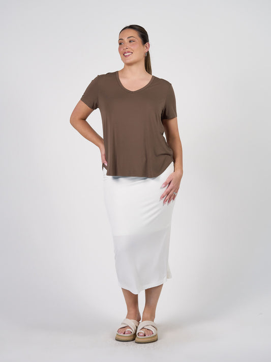 A woman smiles with one hand on her hip, wearing the Veronica Bamboo V-Neck T-Shirt in Chocolate, a white ankle-length skirt, and beige sandals against a plain white background.