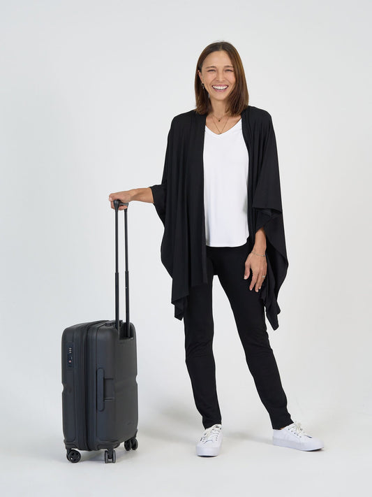 A woman wearing The Long Haul Set—black cardigan, white top, and black pants—stands smiling with one hand on a black rolling suitcase against a plain white background.