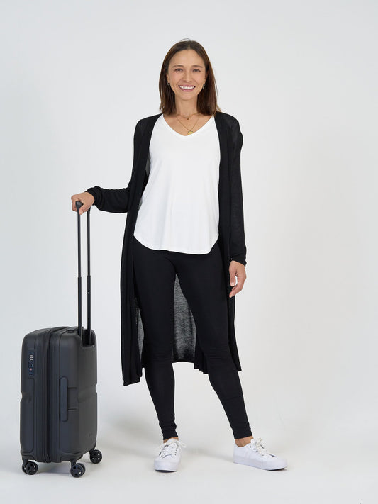 A woman stands and smiles against a white background, wearing The Alpine Set—a long black cardigan, white top, and black leggings—with white sneakers, holding a black rolling suitcase.
