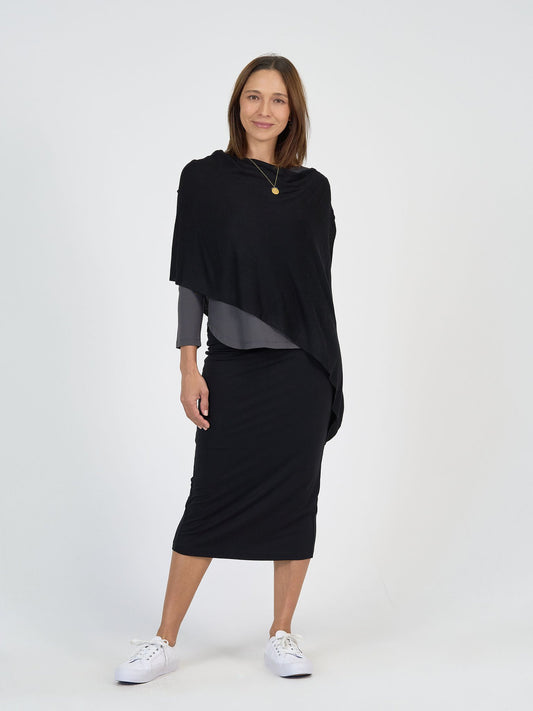 A woman with shoulder-length brown hair wears The Winter Sun Set—featuring a black shawl, gray top, and black midi skirt—paired with white sneakers and a gold necklace, smiling gently against a plain white background.