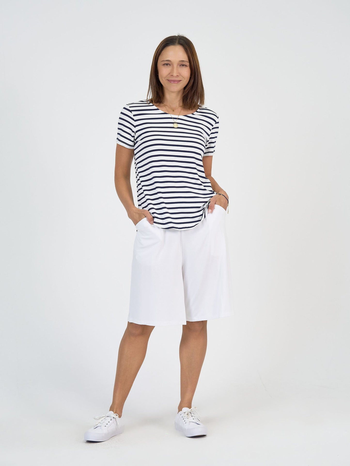 A woman with shoulder-length brown hair smiles gently, standing against a plain white background with her hands in her pockets. She wears The Cruise Set, featuring a black and white striped tee and wide leg bamboo resort shorts, paired with white sneakers.