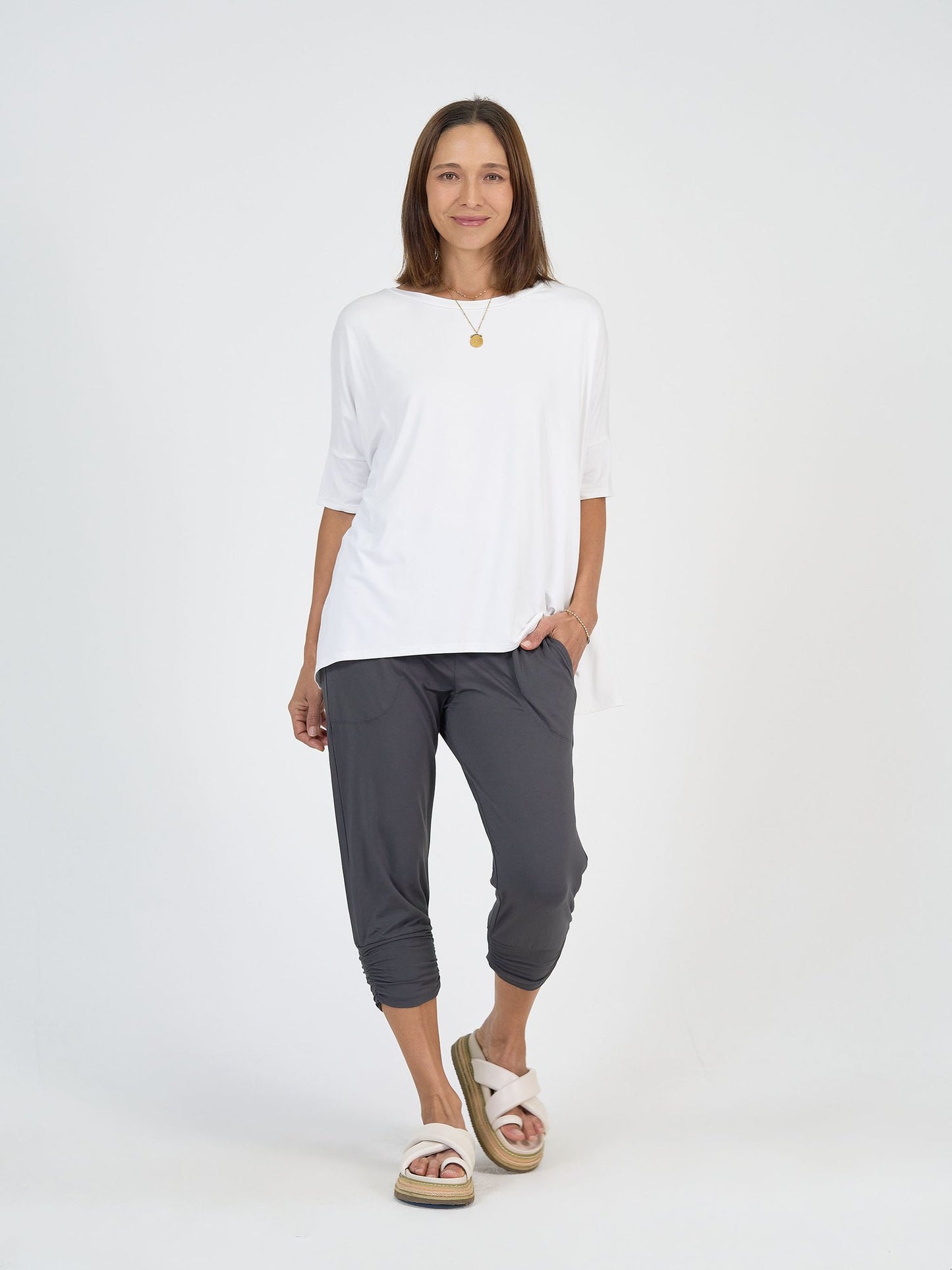 A woman stands against a white background wearing The Long Weekend Set: a loose white T-shirt and gray cropped pants, paired with white slide sandals and a gold pendant necklace, her left hand in her pocket and expression relaxed.