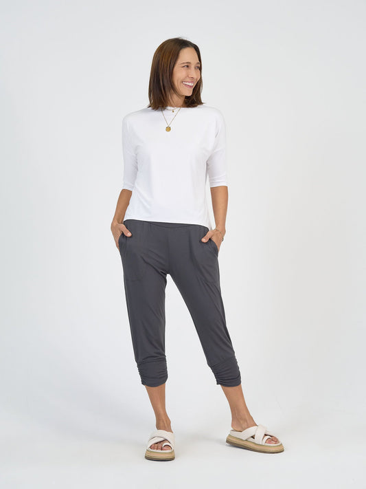 A woman smiles against a white background, wearing The Long Weekend Set: a white three-quarter sleeve top and dark gray cropped jogger pants, styled with white slide sandals and a gold pendant necklace, her hands in her pockets.