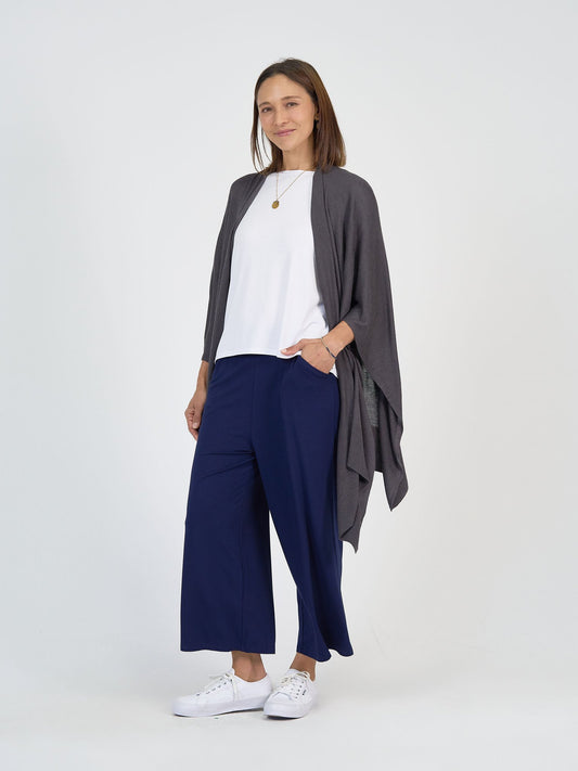 A woman stands against a white background, smiling with one hand in her pocket while wearing The Sea Day Set—featuring a gray draped cardigan, white top, navy wide-leg pants—and white sneakers. She has straight brown hair.