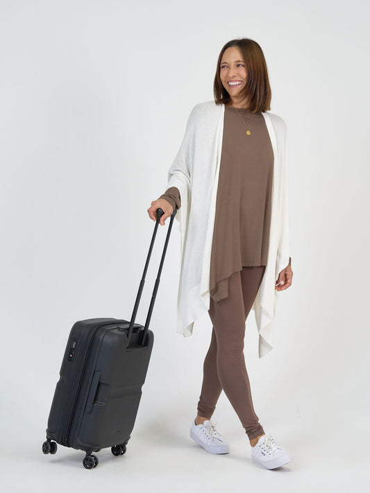 A woman smiles while pulling The Cabin Set, a black rolling suitcase. She wears a brown matching top and leggings, a white open cardigan, and white sneakers—ideal for effortless, packable resort outfits.