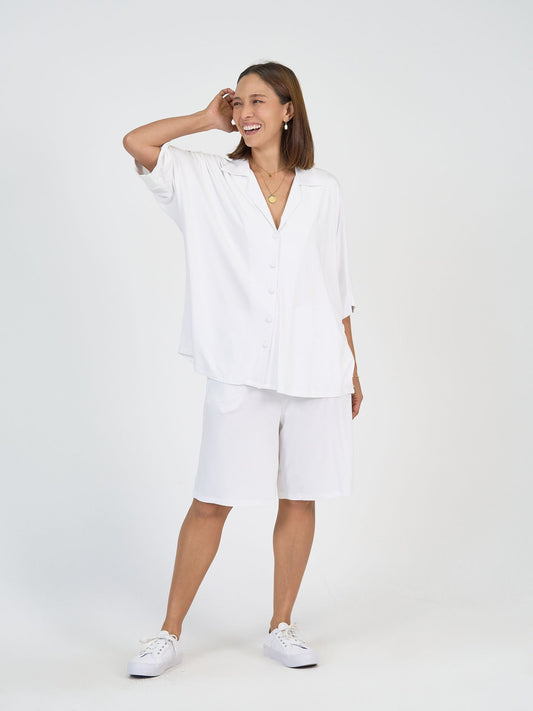 A woman smiles against a plain white background, wearing The Cruise Set with wide leg shorts, a loose-fit shirt, and white sneakers. She accessorizes with gold jewelry and has one hand touching her head.