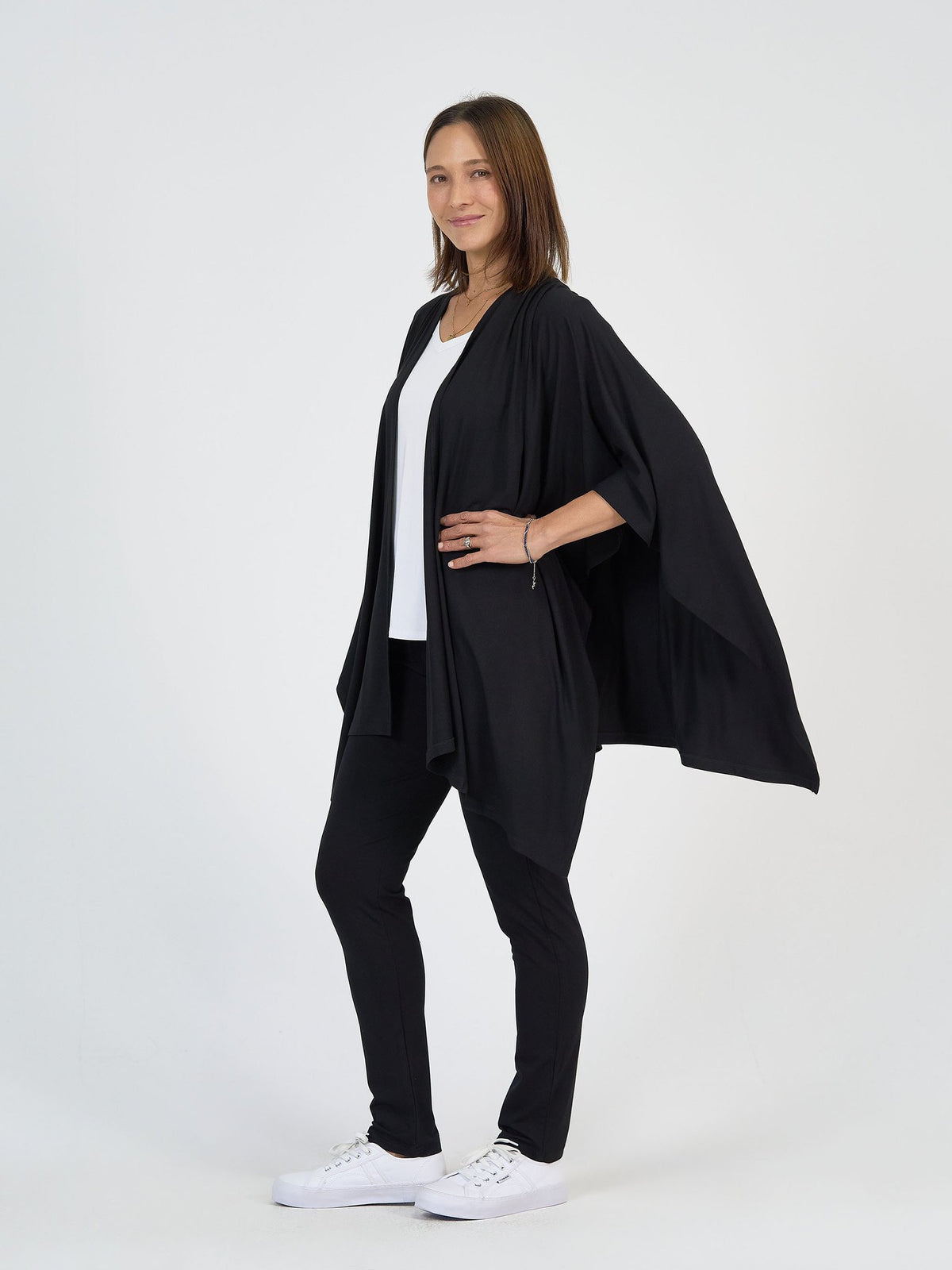 A woman smiles against a white background, posed at an angle with one hand on her hip, wearing The Long Haul Set—a loose black open-front cardigan, white top, black pants—and white sneakers.