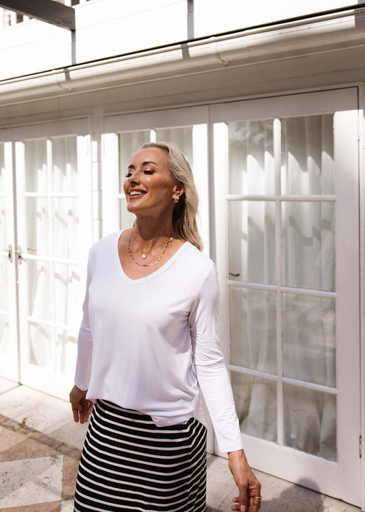A blonde woman smiles outdoors in front of white French doors, wearing the Sia V Neck Bamboo Longsleeve Top in white and a black-and-white striped skirt, as sunlight creates soft shadows around her.