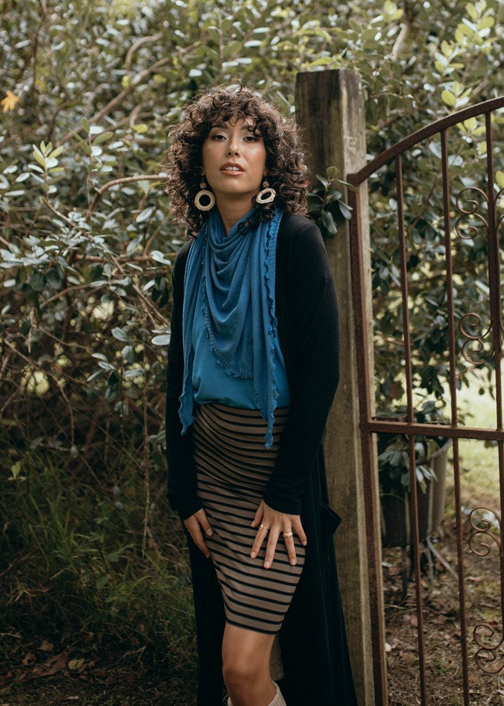 An outdoor shot of a woman wearing a blue scarf, black long line pocket cardigan and a brown striped midi skirt