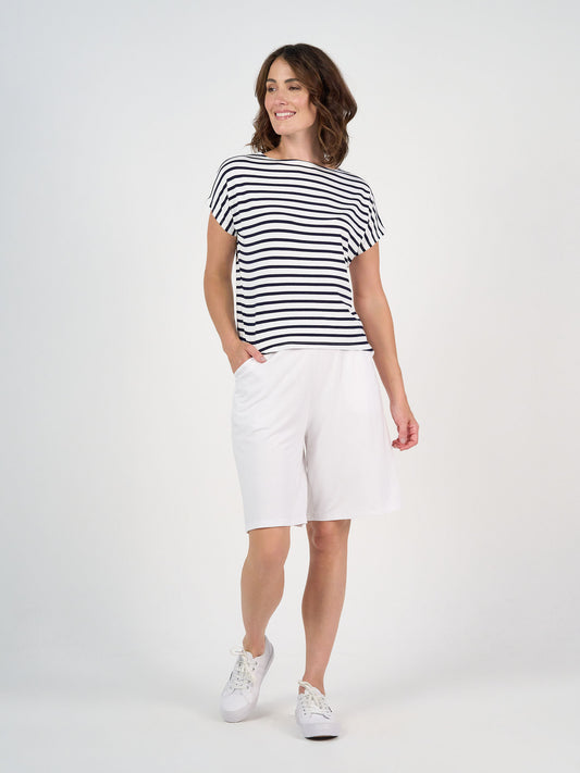 A woman smiles with one hand in her pocket, wearing the Wilde Bamboo Box Fit Tee in navy and white stripe, paired with loose white knee-length shorts and white sneakers against a plain white background.