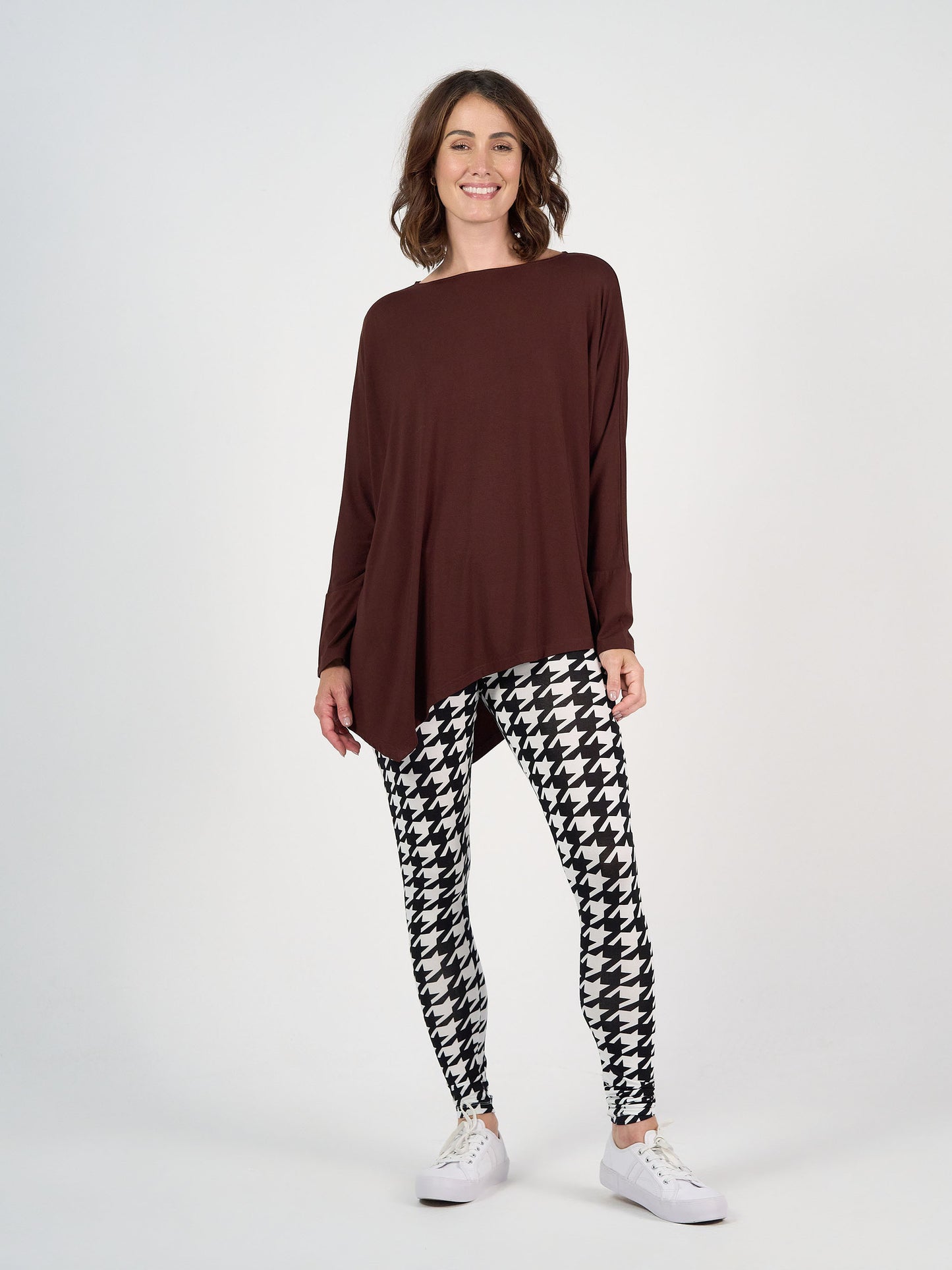 A woman smiles against a plain background, wearing the Susie Oversized Asymmetrical Bamboo Top in Burgundy, paired with black and white houndstooth leggings and white sneakers.