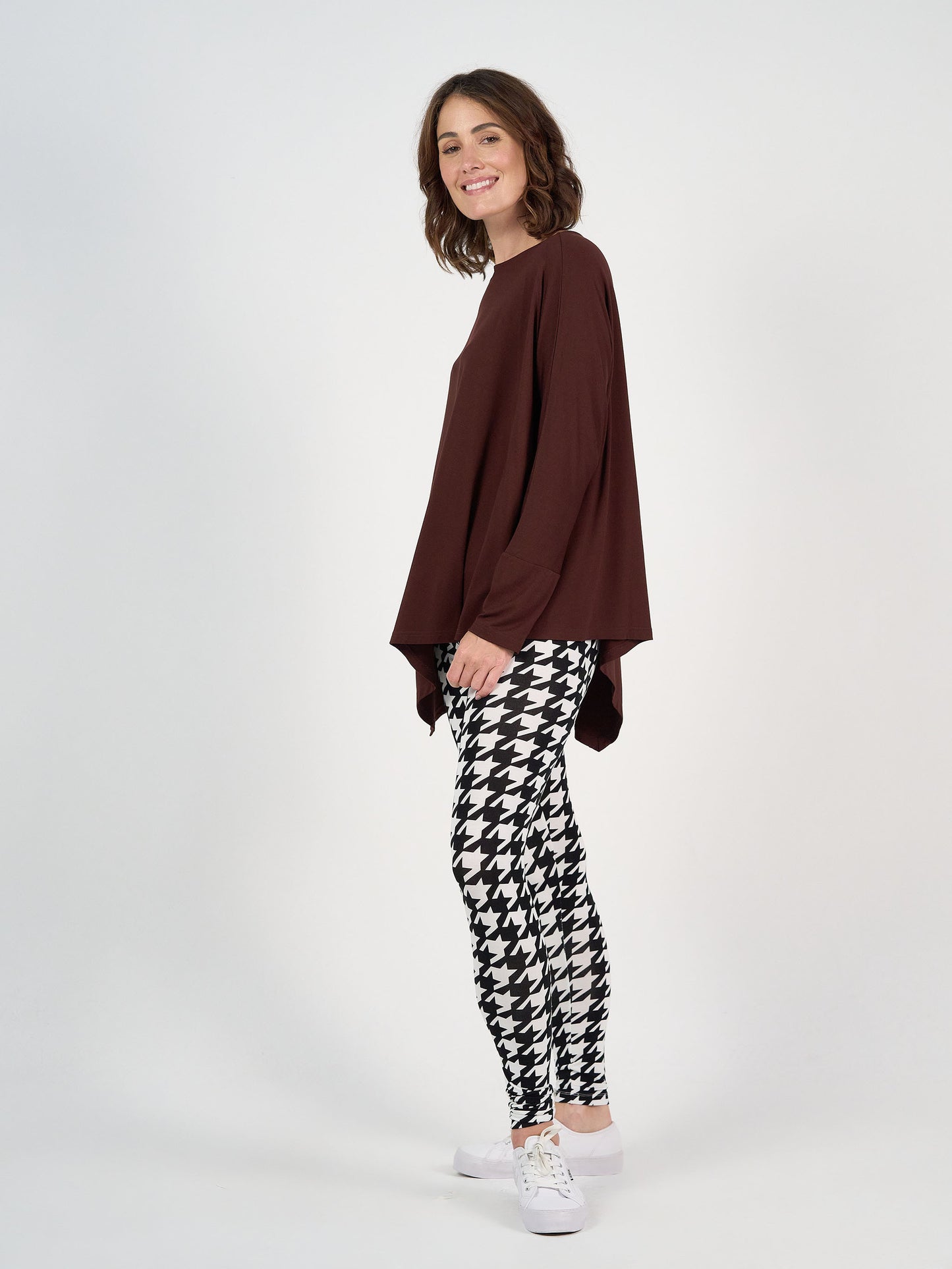 A woman stands sideways, smiling, wearing the Susie Oversized Asymmetrical Bamboo Top in burgundy, paired with black and white houndstooth leggings and white sneakers against a plain white background.