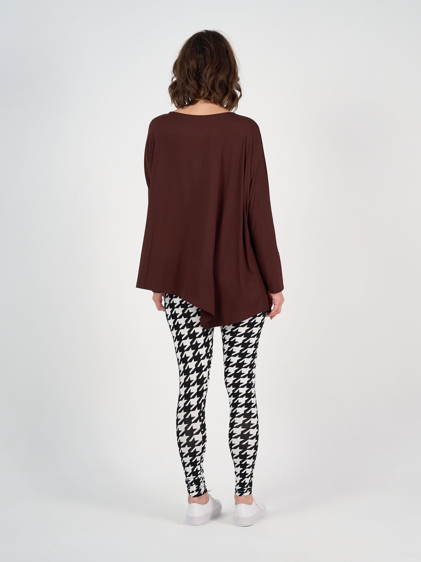 A person stands with their back to the camera, wearing the Susie Oversized Asymmetrical Bamboo Top in burgundy, black and white houndstooth leggings, and white shoes against a plain white background.