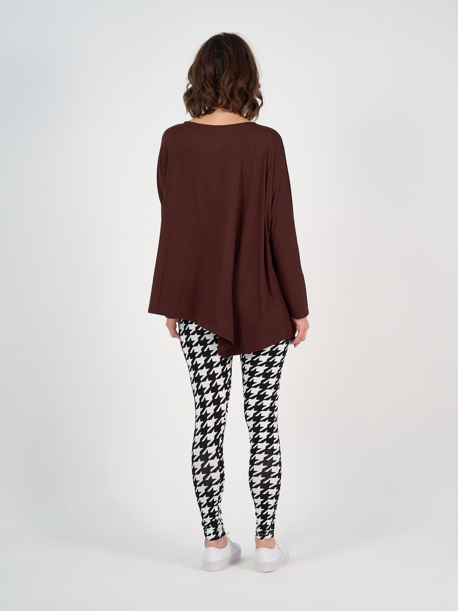 A person stands with their back to the camera, wearing the Susie Oversized Asymmetrical Bamboo Top in burgundy, black and white houndstooth leggings, and white shoes against a plain white background.