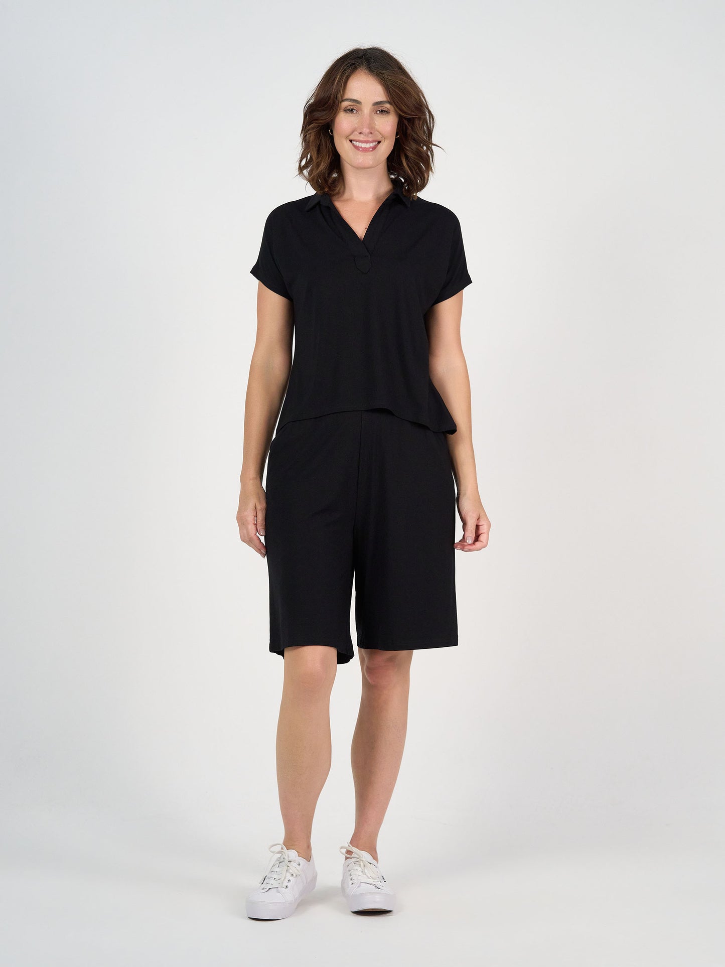 A woman stands smiling against a plain white background, wearing the Rush Bamboo Short Sleeve Collared Polo in black paired with matching knee-length shorts and white sneakers.