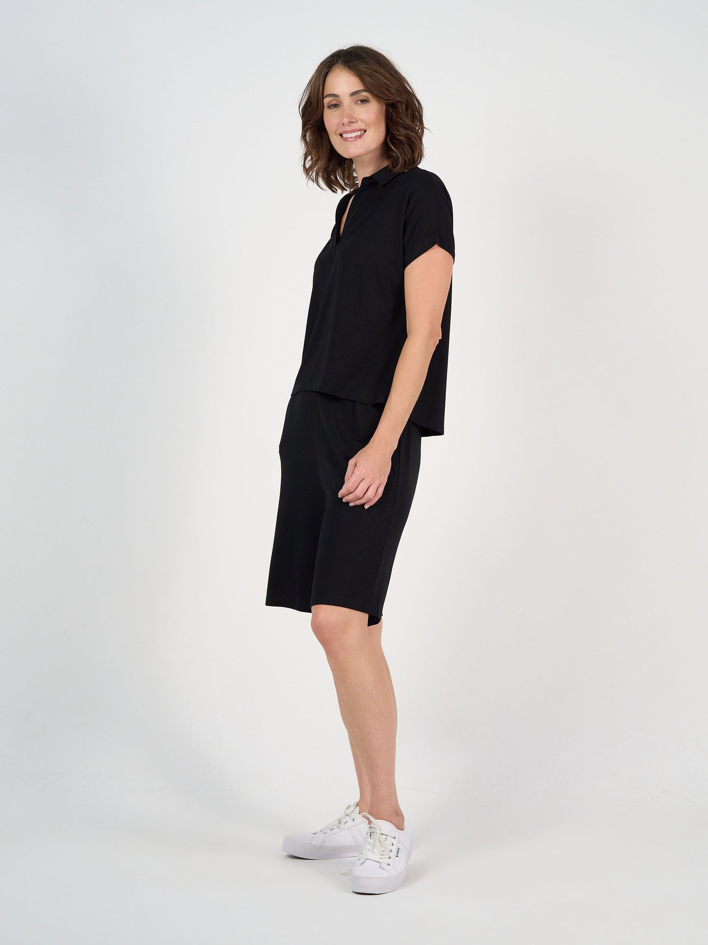 A woman with shoulder-length brown hair smiles sideways, wearing the Rush Bamboo Short Sleeve Collared Polo in black with matching knee-length shorts and white sneakers, against a plain white background.