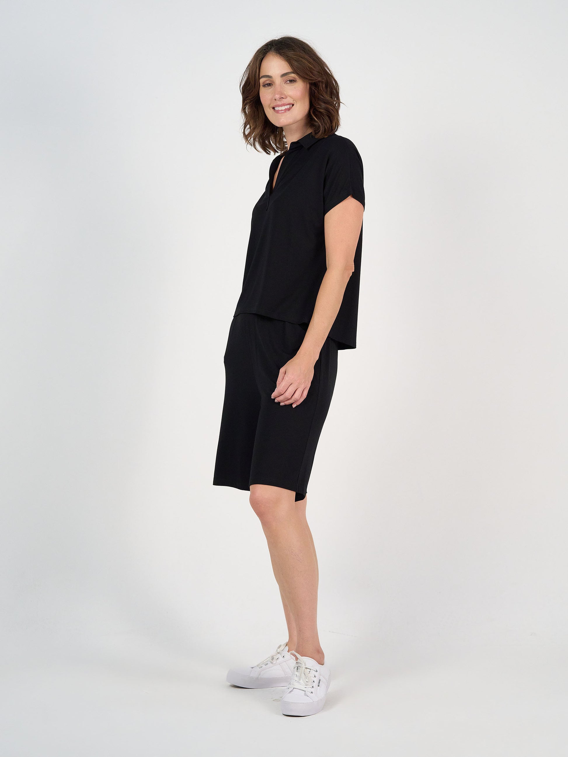 A woman with shoulder-length brown hair smiles sideways, wearing the Rush Bamboo Short Sleeve Collared Polo in black with matching knee-length shorts and white sneakers, against a plain white background.