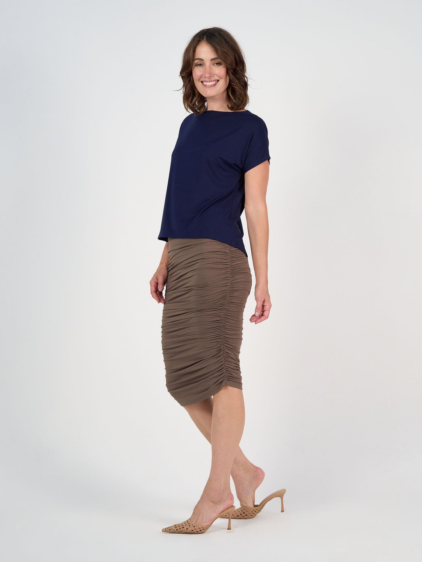 A woman smiles in the Wilde Bamboo Box Fit Tee in navy, paired with a taupe ruched midi skirt and beige heeled mules, set against a plain white background.