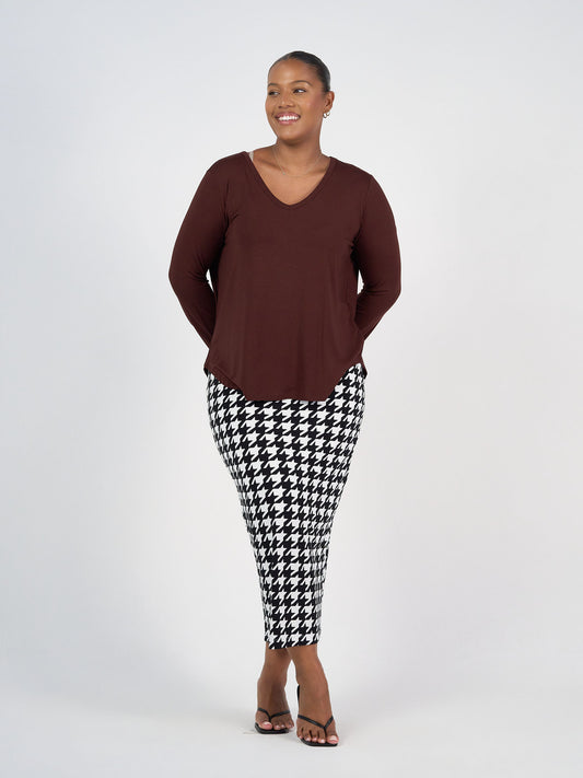 A woman smiles against a plain white background, wearing the Sia V Neck Bamboo Longsleeve Top in Burgundy with black-and-white houndstooth pants and black sandals. Her hands are behind her back—an ideal soft layering piece for any season.