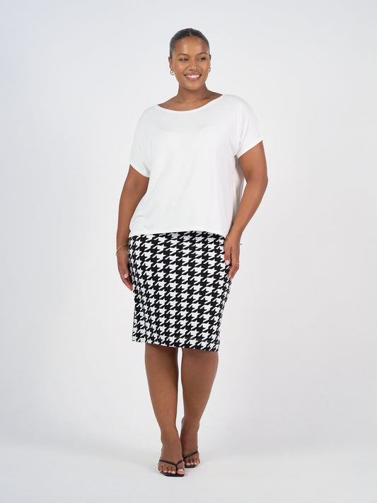 A woman smiles against a white background, wearing a loose white short-sleeve top, black sandals, and the Midi Bamboo Whitney Tube Skirt in houndstooth with a stretch waistband.