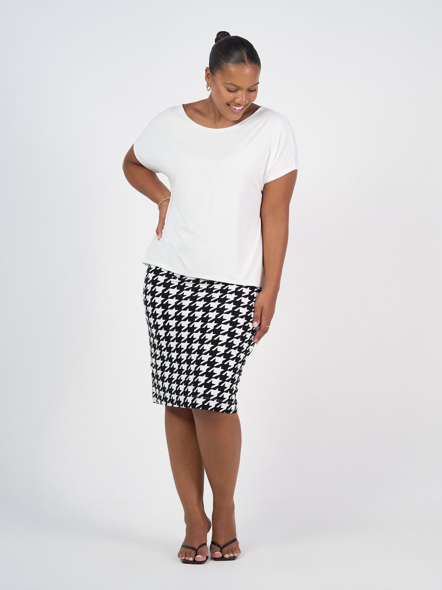A woman stands smiling, wearing the Wilde Bamboo Box Fit Tee in cream with a black-and-white houndstooth pencil skirt and black sandals, against a plain white background.