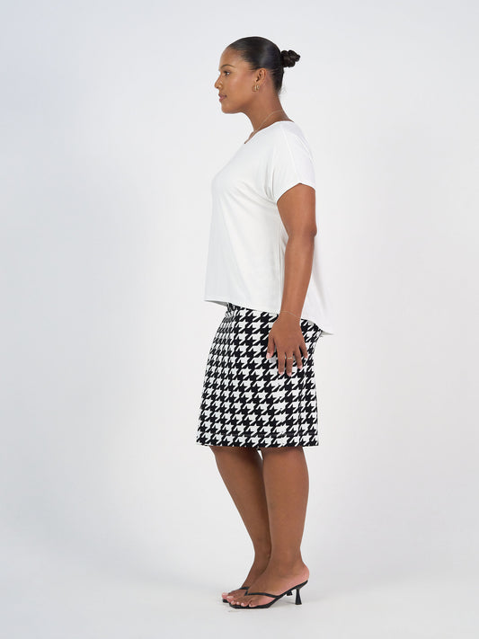 A woman in profile against a white background wears the Wilde Bamboo Box Fit Tee in cream, paired with a black and white houndstooth skirt and black heeled sandals. Her hair is styled in a neat bun.