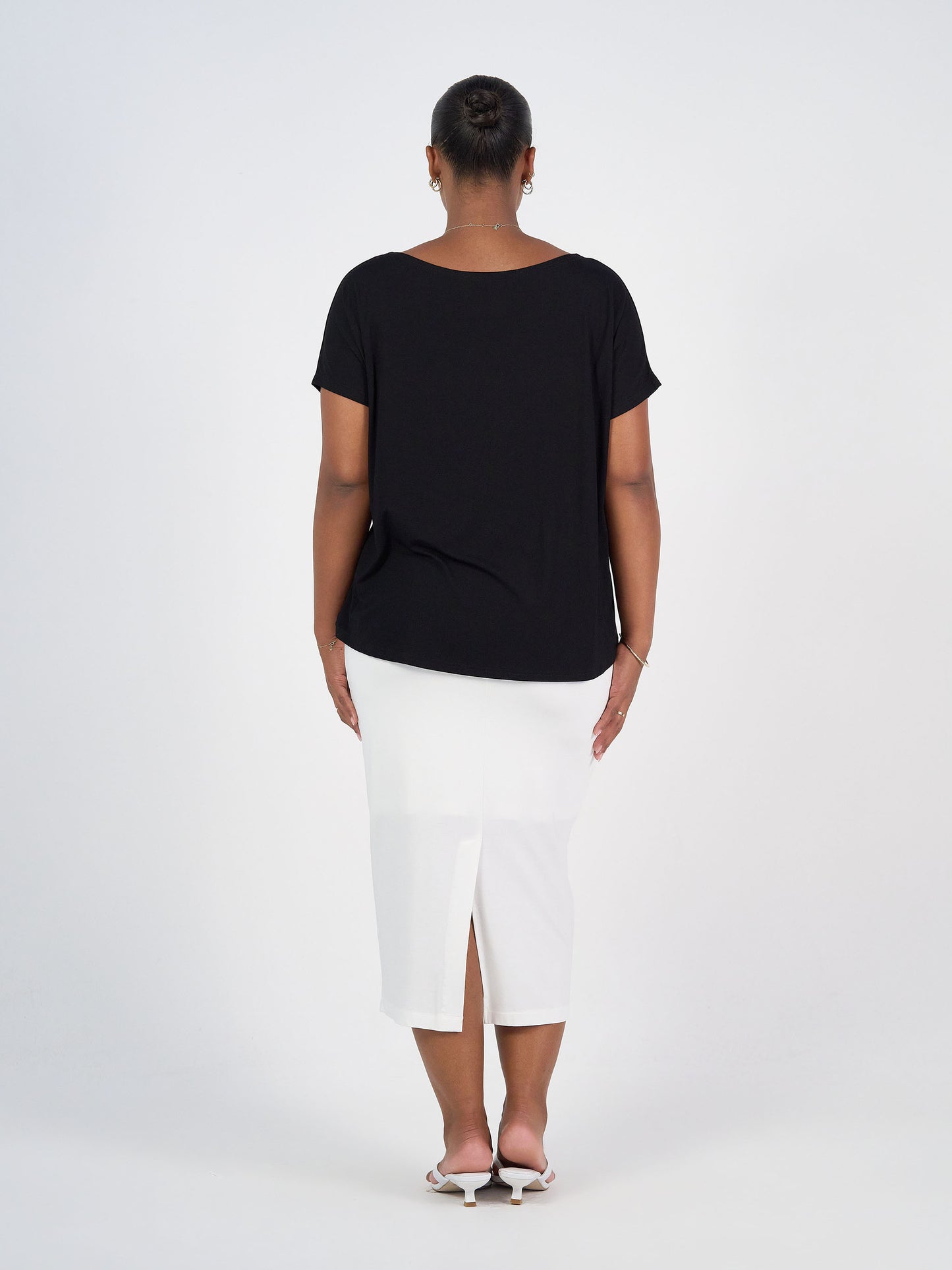 A woman stands with her back to the camera, wearing the Wilde Bamboo Box Fit Tee in black, a white knee-length skirt with a back slit, and open-toe heels against a plain white background.