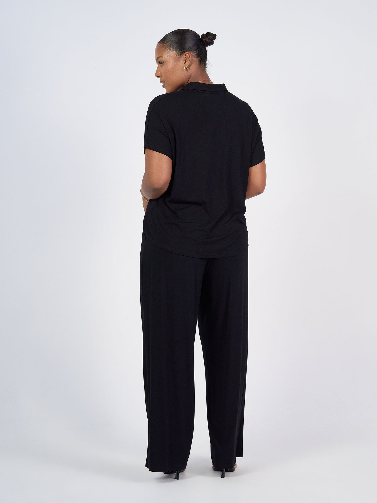 A woman stands with her back to the camera, wearing the Rush Bamboo Short Sleeve Collared Polo in black and matching pants. Her hair is styled in a neat bun as she poses against a plain white background.