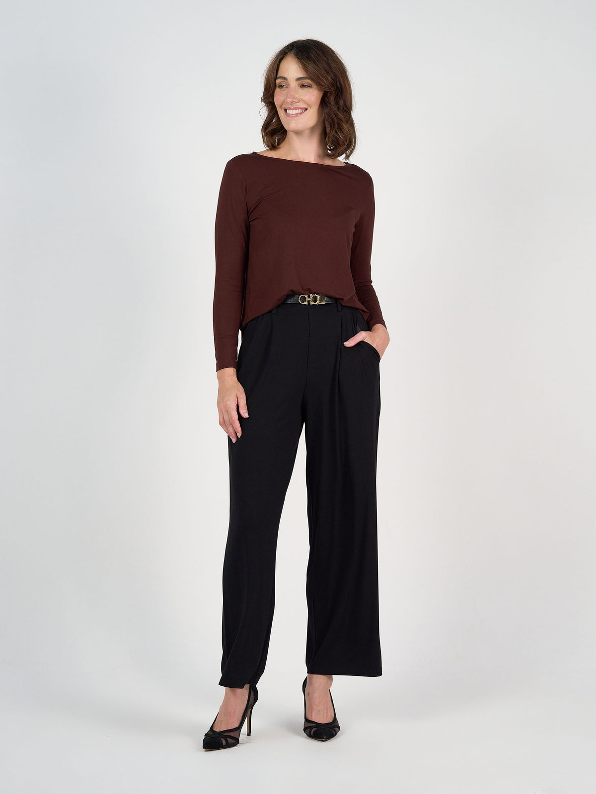 A woman stands smiling against a plain background, wearing the Adele Long Sleeve Bamboo Top in Burgundy, black high-waisted belted pants, and black heels. Her left hand is in her pocket as she looks slightly to the right.