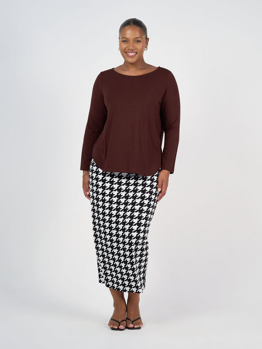 A woman stands smiling, wearing the Adele Long Sleeve Bamboo Top in burgundy, a black-and-white houndstooth skirt, and black sandals against a plain light background.