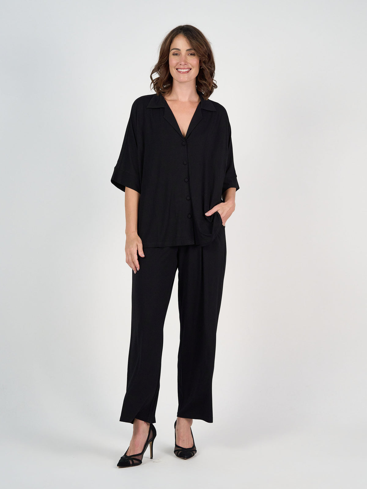 A woman stands against a white background, smiling and radiating confidence. She wears the Alanis 3/4 Sleeve Collared Bamboo Shirt in black with matching pants and heels, her hand in her pocket for a relaxed look.