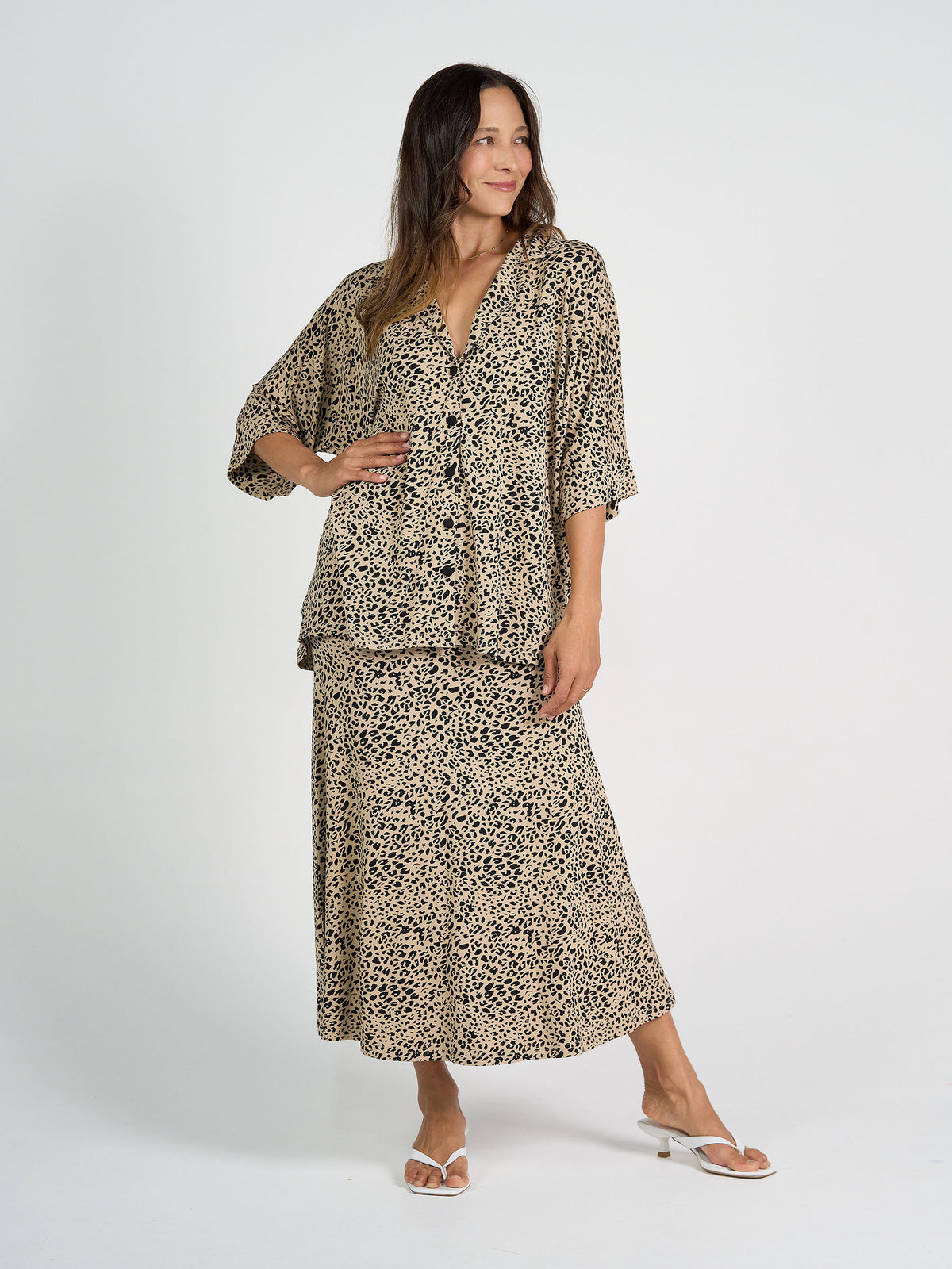 A woman stands against a white background wearing the Alanis 3/4 Sleeve Collared Shirt in brown leopard print with matching midi skirt and white sandals. She smiles, her long hair down, one hand on her hip to show the flowy silhouette.