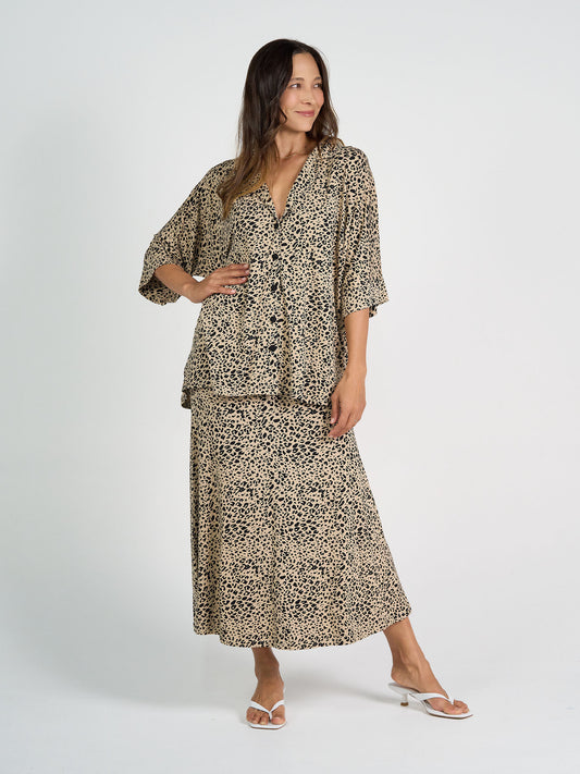 A woman stands against a white background wearing the Alanis 3/4 Sleeve Collared Shirt in brown leopard print with matching midi skirt and white sandals. She smiles, her long hair down, one hand on her hip to show the flowy silhouette.