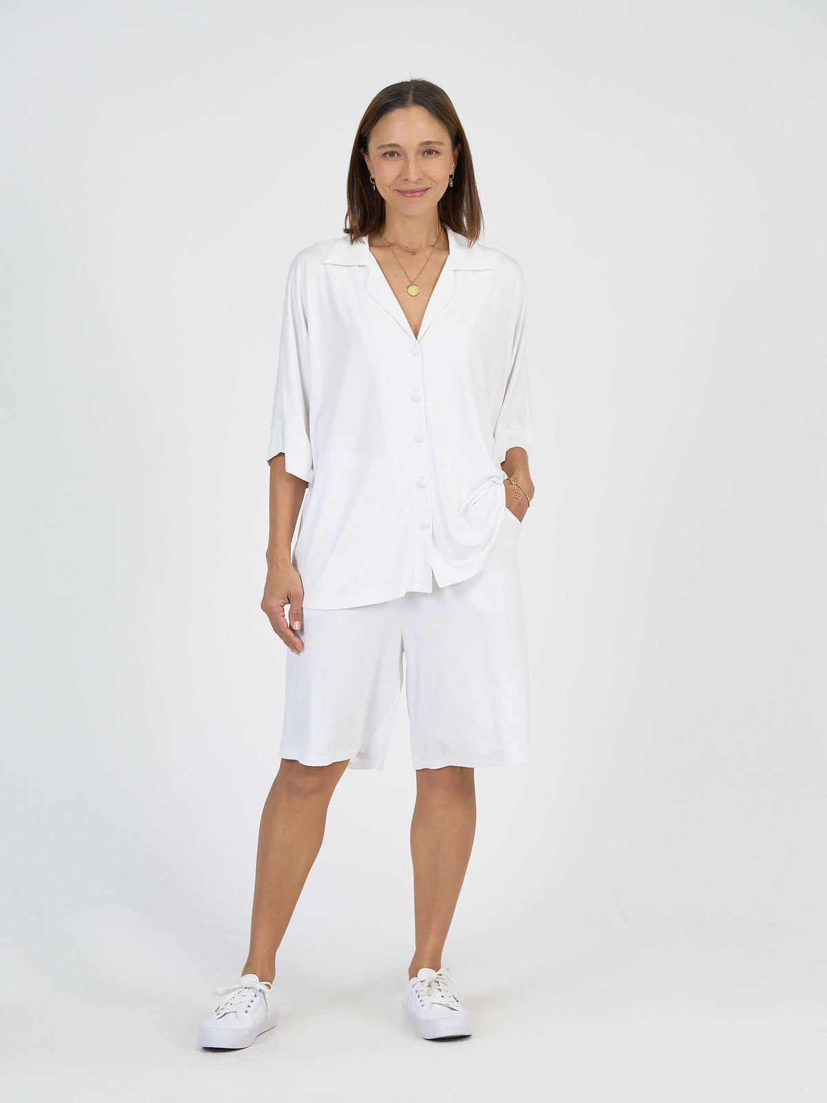 A woman stands against a plain white background wearing a loose white button-up shirt, white knee-length shorts, white sneakers, and two gold necklaces. She has straight brown hair and is smiling gently with one hand in her pocket.