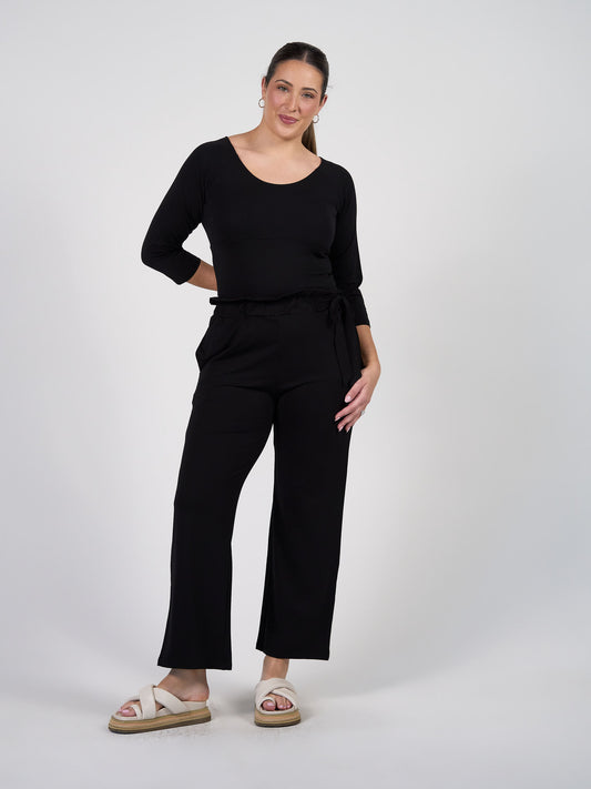 A woman poses against a white background, smiling with one hand on her hip. She wears the Ava 3/4 Sleeve Cropped Bamboo Top in black, styled with black wide-leg pants and white sandals—an ideal layering piece.