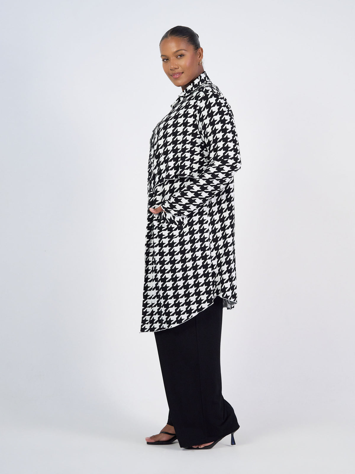A woman stands sideways in the Avril Bamboo Trench Coat - Houndstooth over black pants and open-toe heels. She smiles gently, hands in pockets, against a plain white background—blending style and sustainability.
