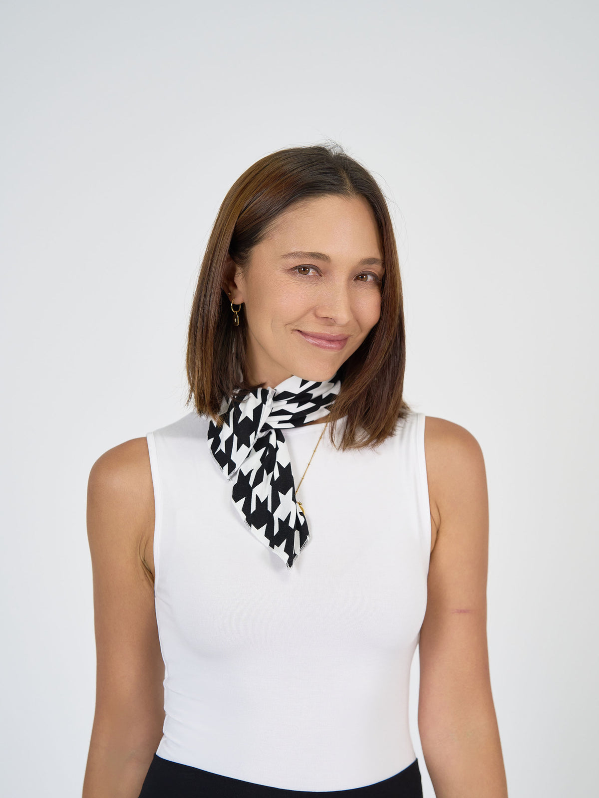 A woman with straight brown hair wears a white sleeveless top and the Benni Bamboo Scarf - Houndstooth around her neck, smiling softly against a plain white background.