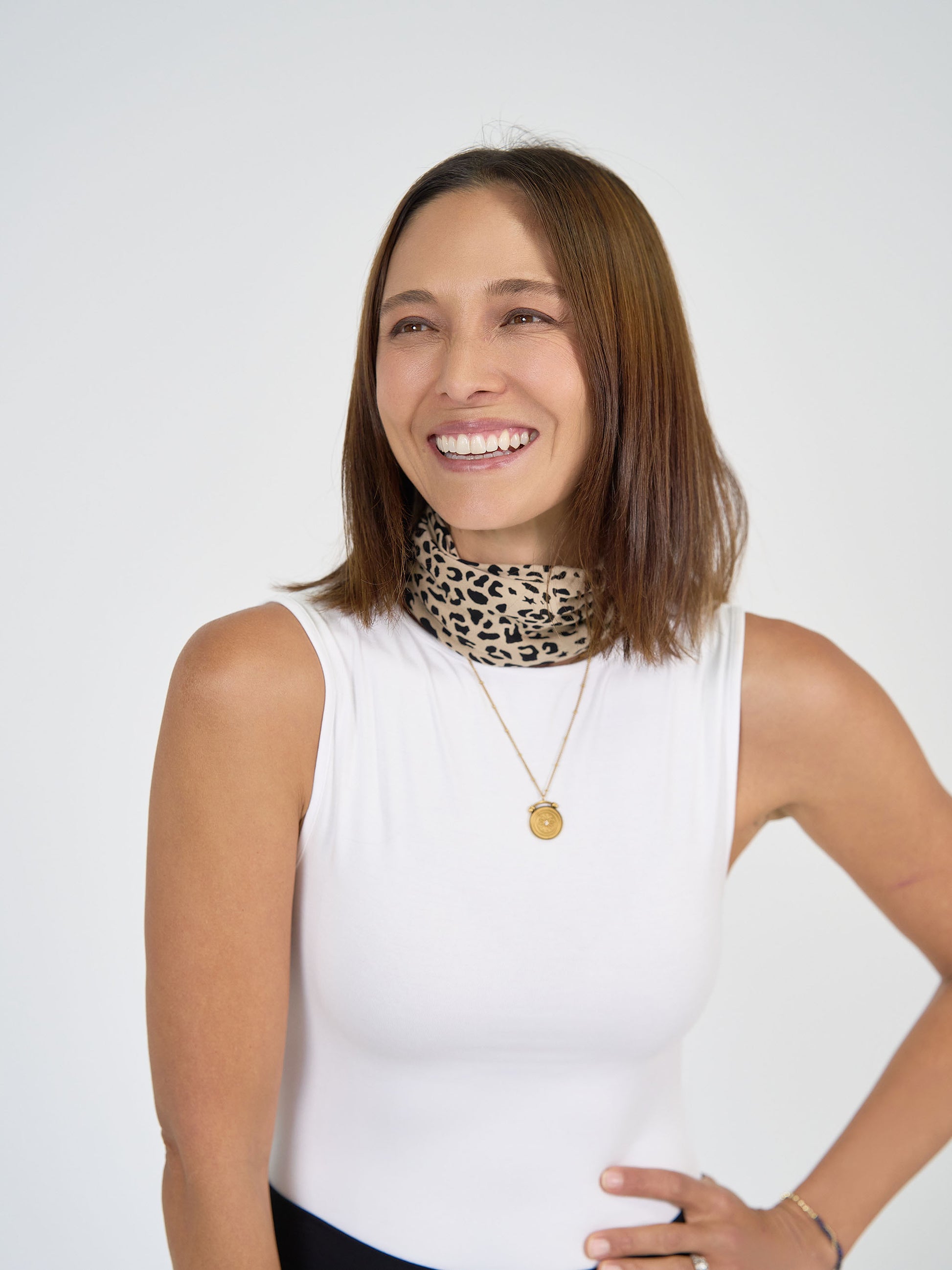 A woman with straight brown hair wears a white sleeveless top, a gold necklace, and the Benni Bamboo Scarf - Brown Leopard as an accessory. She smiles while looking to the side against a plain light background.