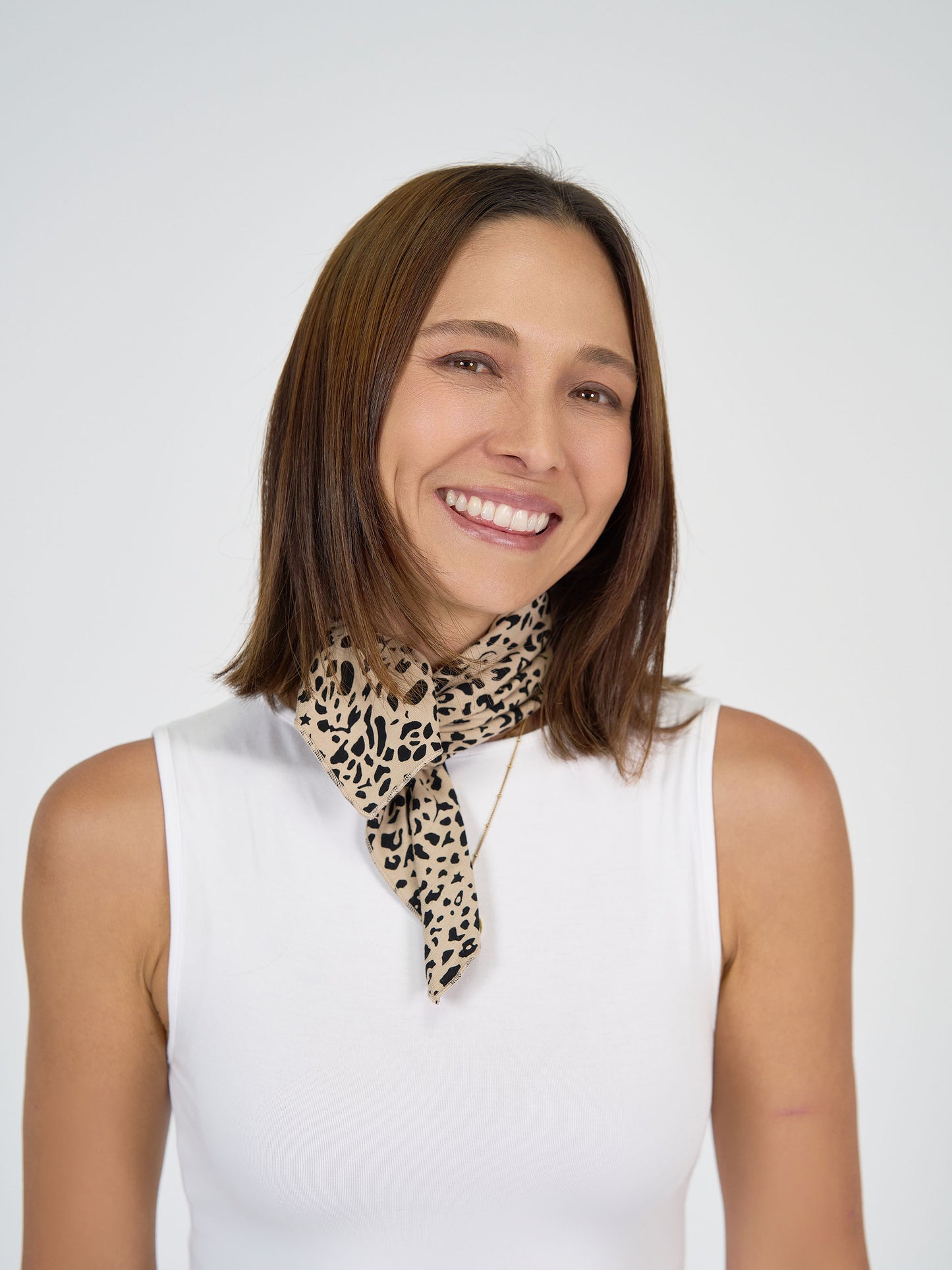 A woman with straight brown hair, wearing a white sleeveless top and the Benni Bamboo Scarf - Brown Leopard, smiles against a plain white background, showcasing this sustainable leopard-print accessory's chic styling.