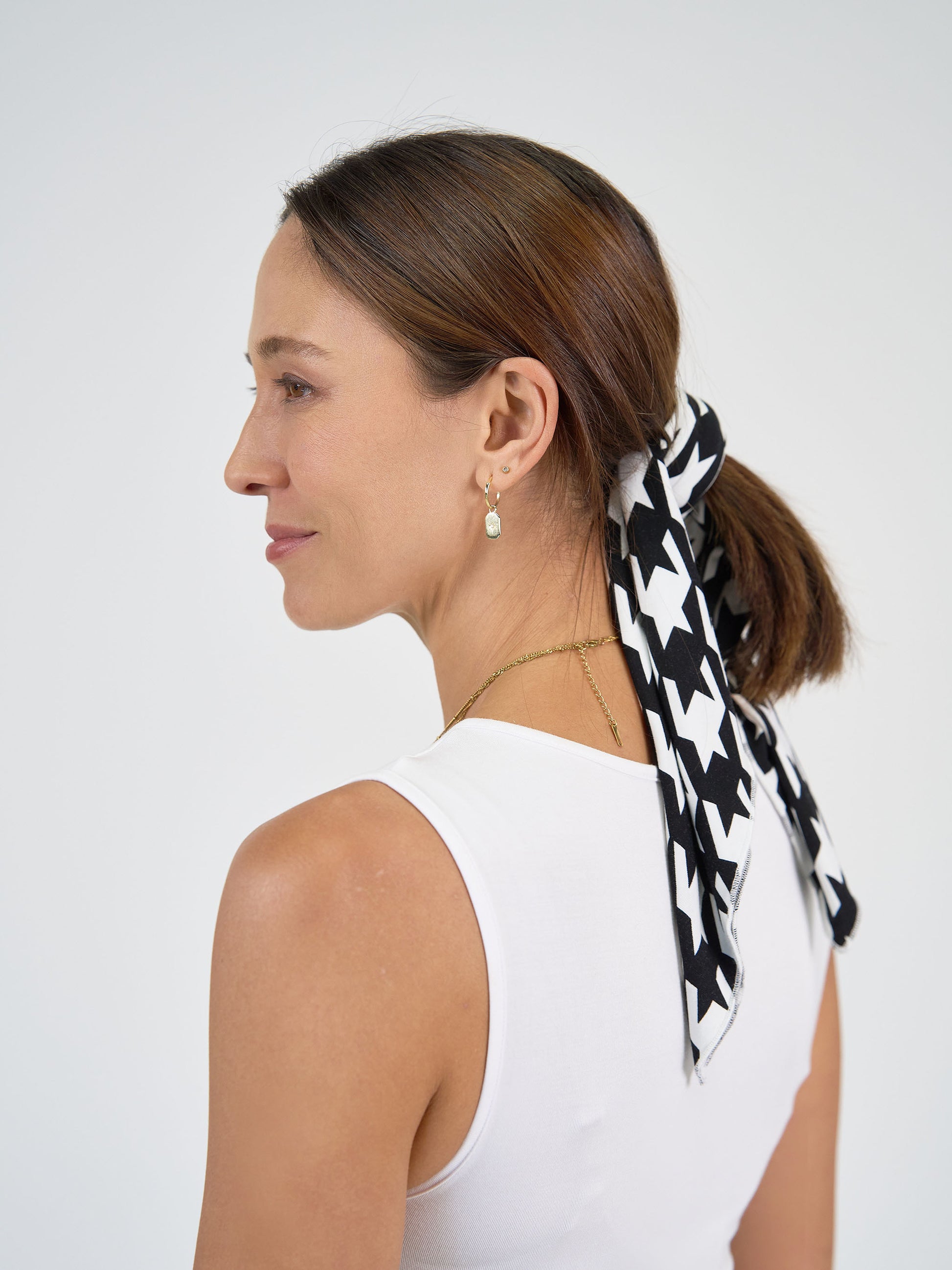 A woman with straight brown hair tied back with the Benni Bamboo Scarf - Houndstooth, wearing a white sleeveless top, gold necklace, and small gold earrings, faces sideways against a plain light background.
