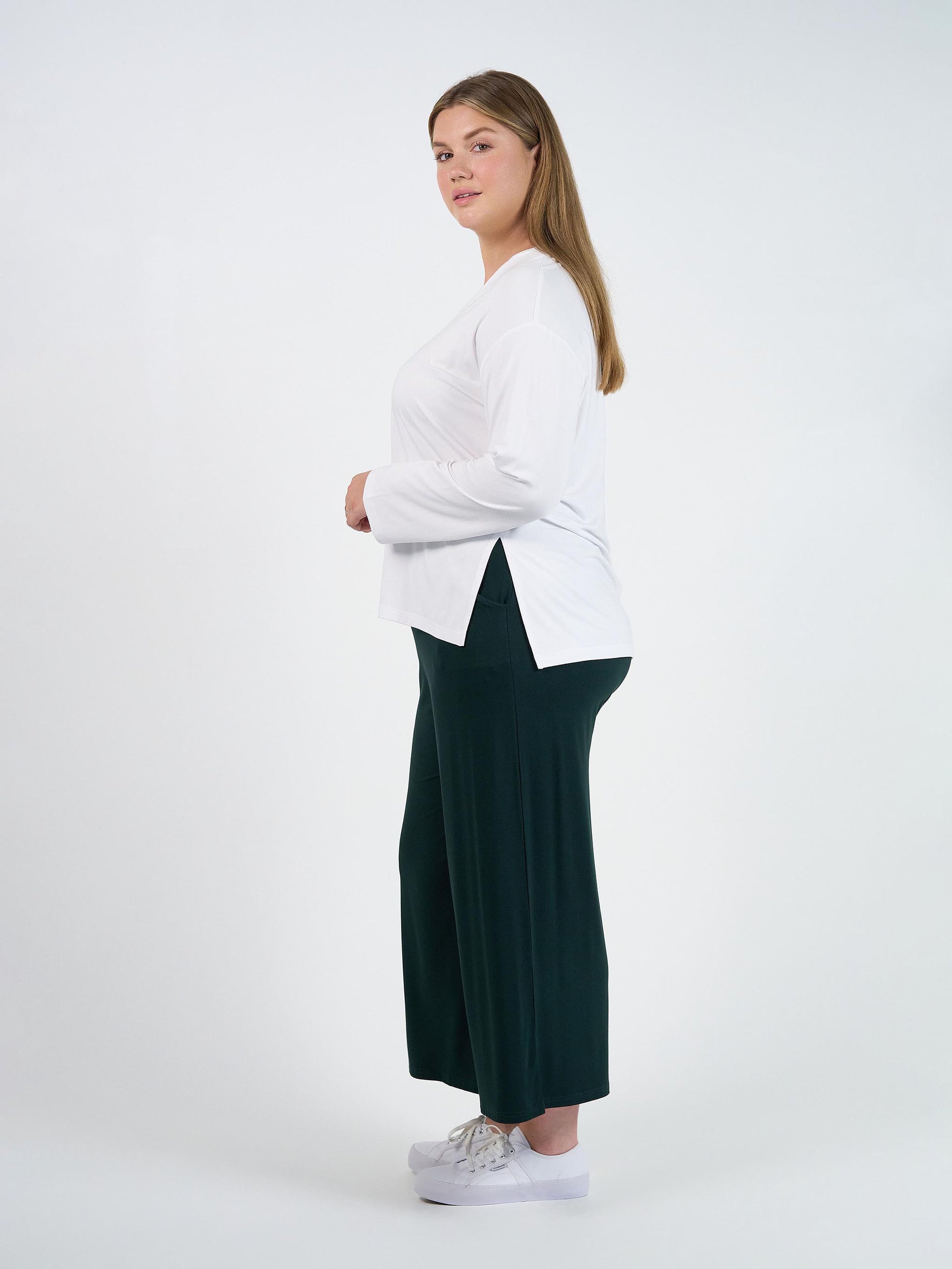 A woman with long brown hair stands sideways against a white background, wearing the Braxton Bamboo Long Sleeve Top in white, loose dark green pants, and white sneakers.