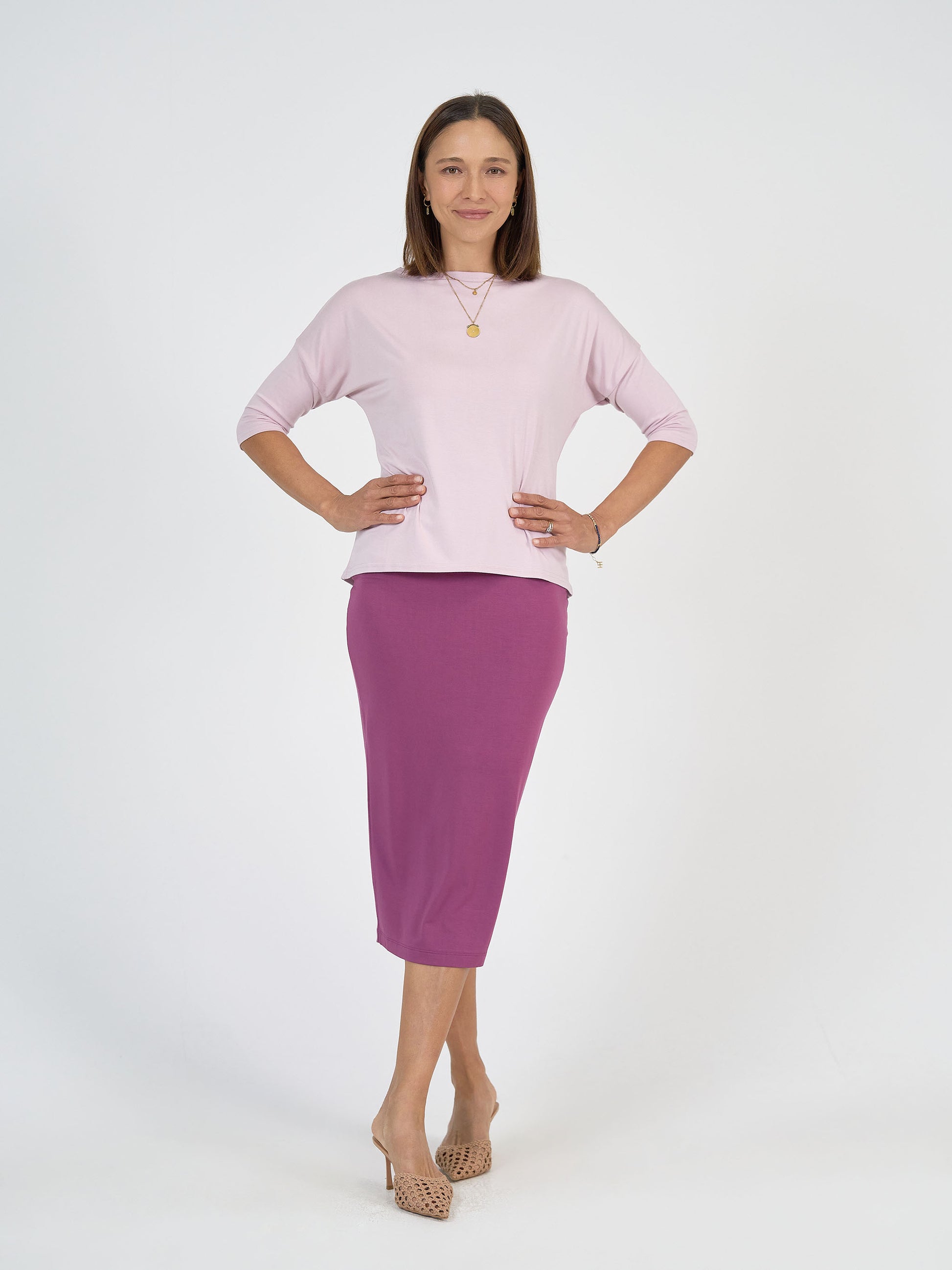 A woman stands confidently in the Maxi Bamboo Whitney Tube Skirt in plum, paired with a light pink top, beige open-toe heels, and a gold necklace, with her hands on her hips against a plain white background.
