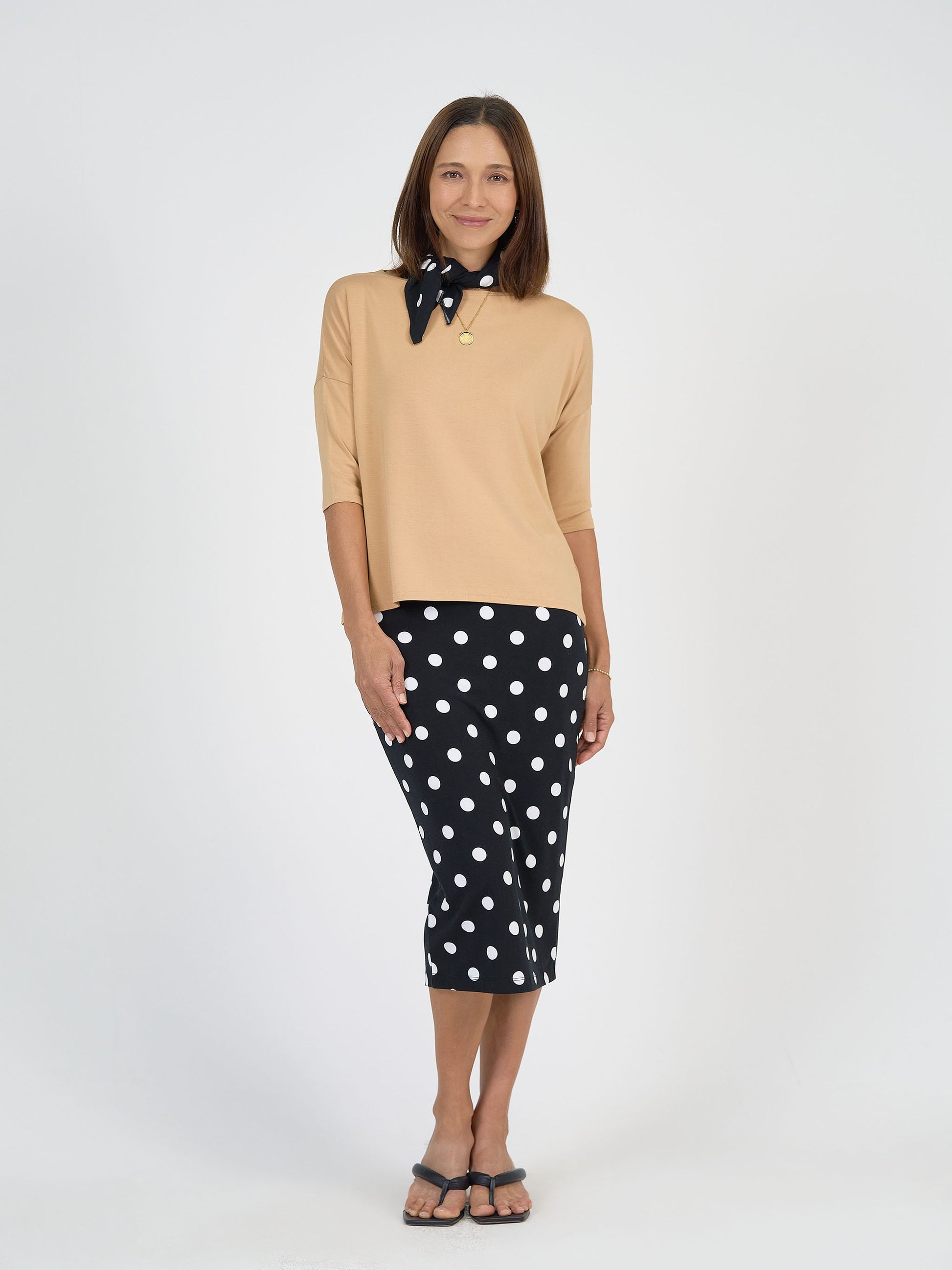A woman smiles against a plain background, wearing a beige top, sandals, a gold necklace, a black midi skirt with white polka dots, and the Benni Bamboo Scarf - Polka Dot styled around her neck—a chic look with this sustainable accessory.