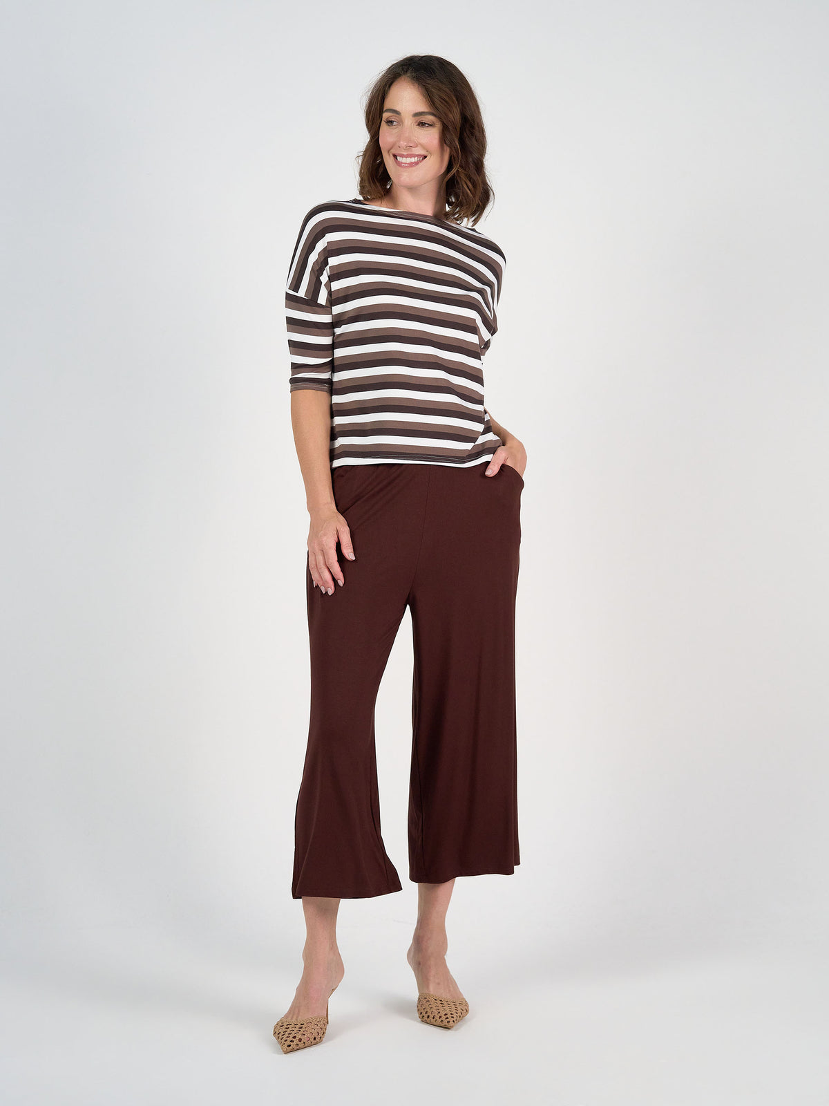 A woman smiles in the Camila 3/4 Sleeve Bamboo Top—brown and white stripes with a relaxed box cut—paired with loose brown cropped pants and beige woven flats, posed against a plain white background.