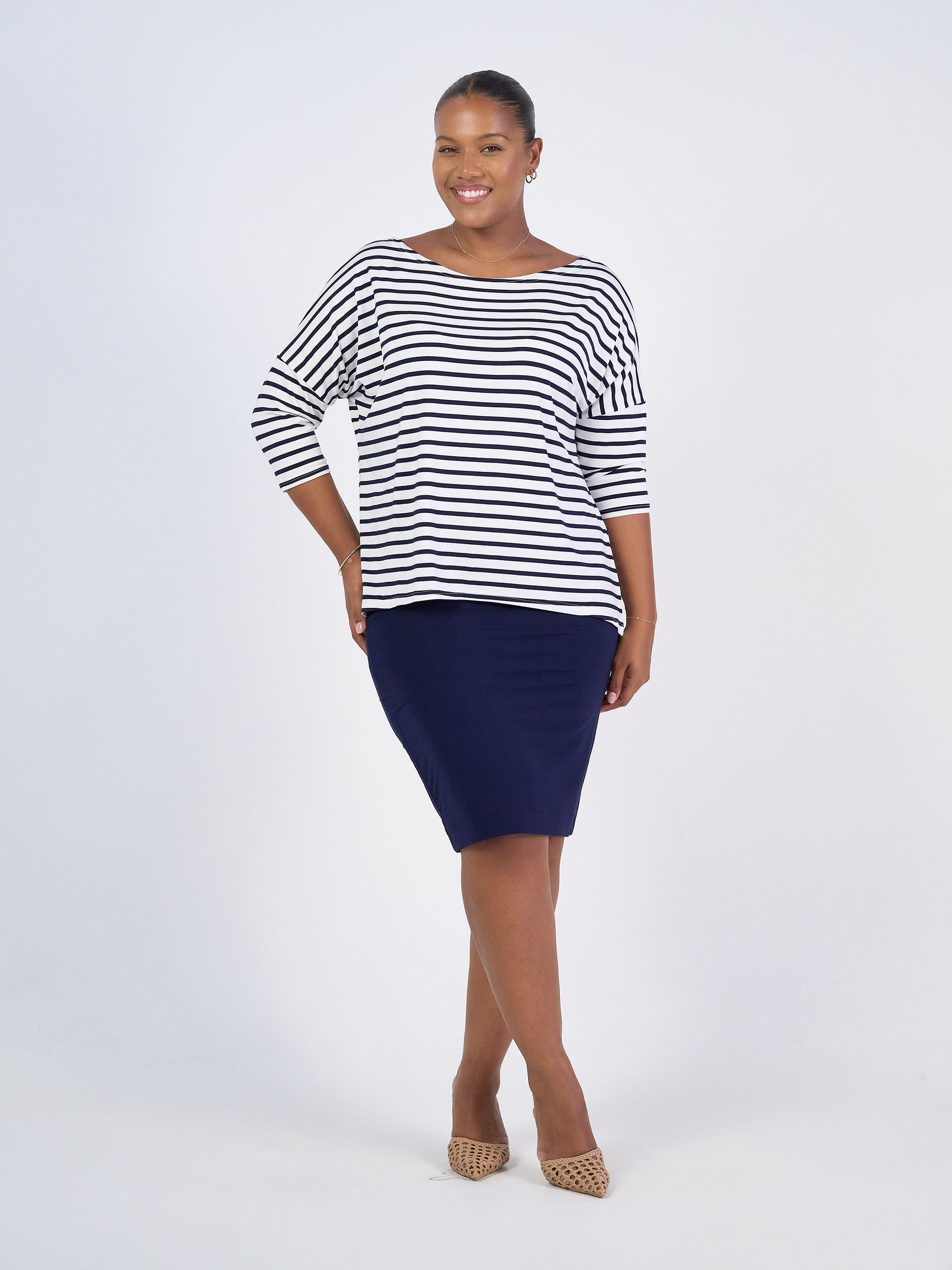 A woman smiles against a plain white background, wearing the Camila 3/4 Sleeve Bamboo Top in black-and-white stripes, paired with a navy knee-length skirt and tan heels.
