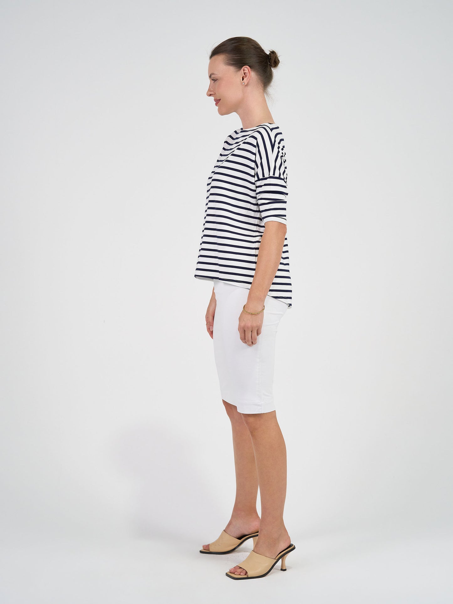A woman in profile stands against a white background, wearing the Camila 3/4 Sleeve Bamboo Top in navy & white stripe, paired with a white knee-length skirt and beige heeled sandals. Her hair is styled in a neat bun.