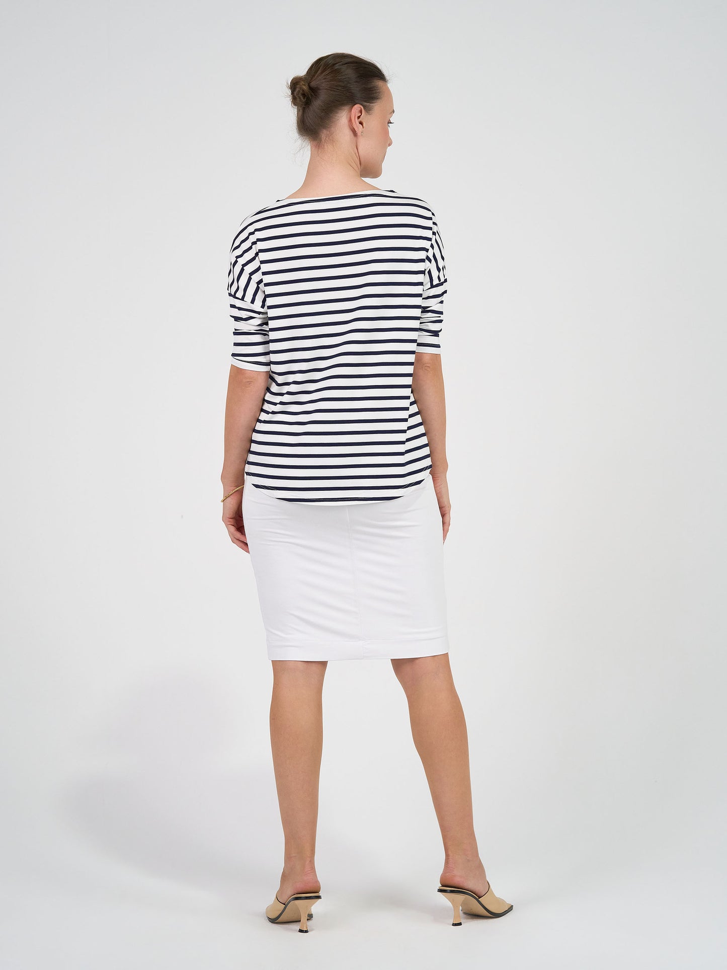 A woman stands with her back to the camera, wearing the Camila 3/4 Sleeve Bamboo Top in navy and white stripe, paired with a white knee-length skirt and beige heels, set against a plain white background.