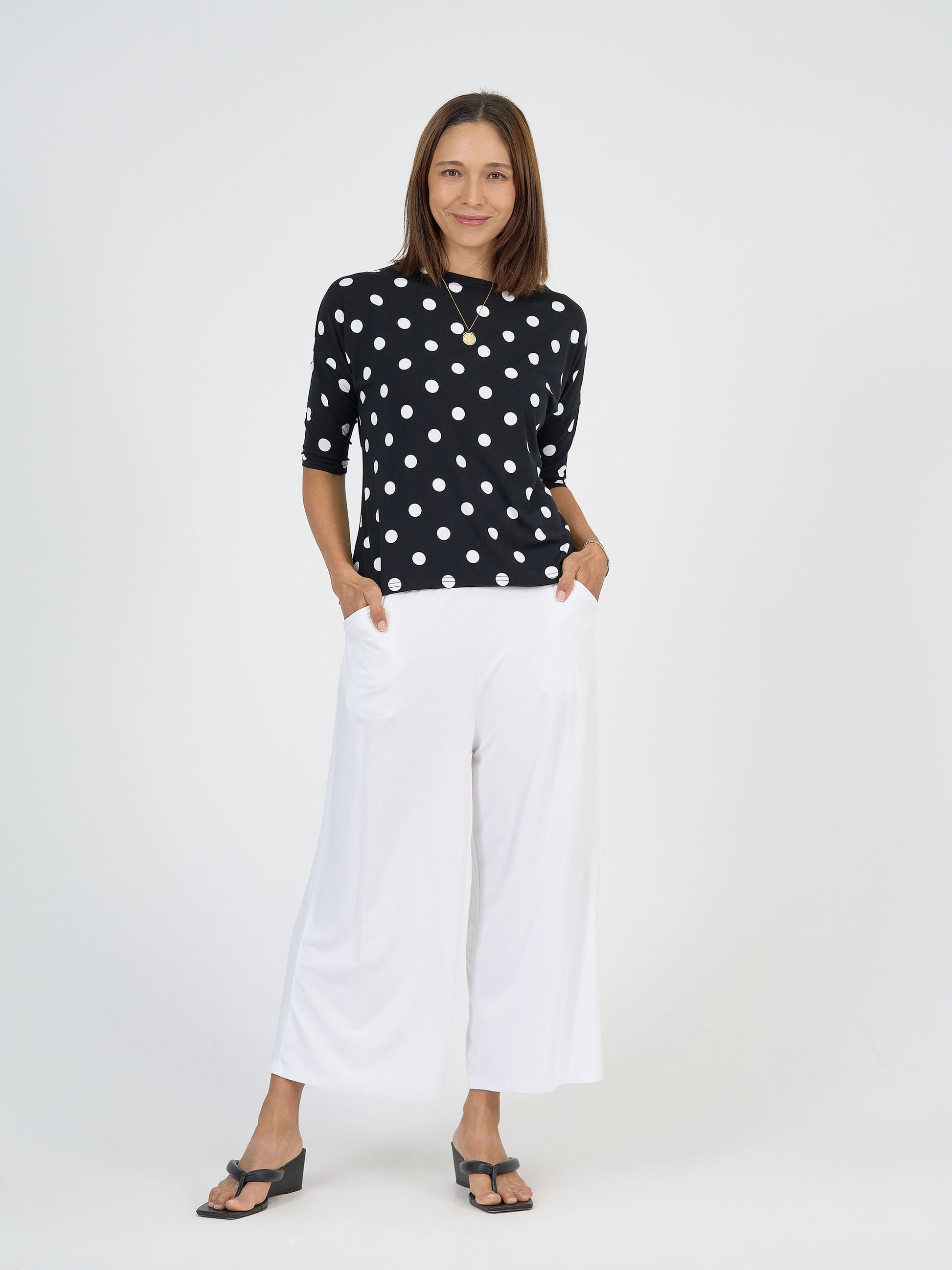 A woman stands against a white background, smiling slightly with her hands in her pockets, wearing the Camila 3/4 Sleeve Bamboo Top - Polka Dot, wide-legged white pants, black sandals, and a necklace.