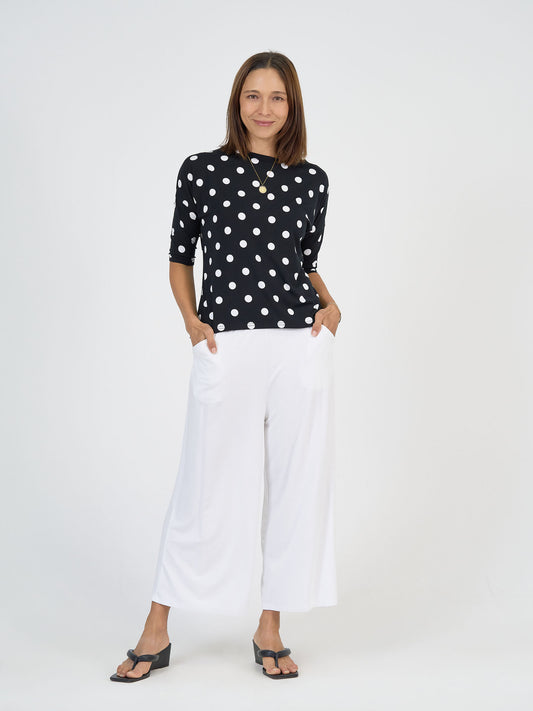 A woman stands against a white background, smiling slightly with her hands in her pockets, wearing the Camila 3/4 Sleeve Bamboo Top - Polka Dot, wide-legged white pants, black sandals, and a necklace.
