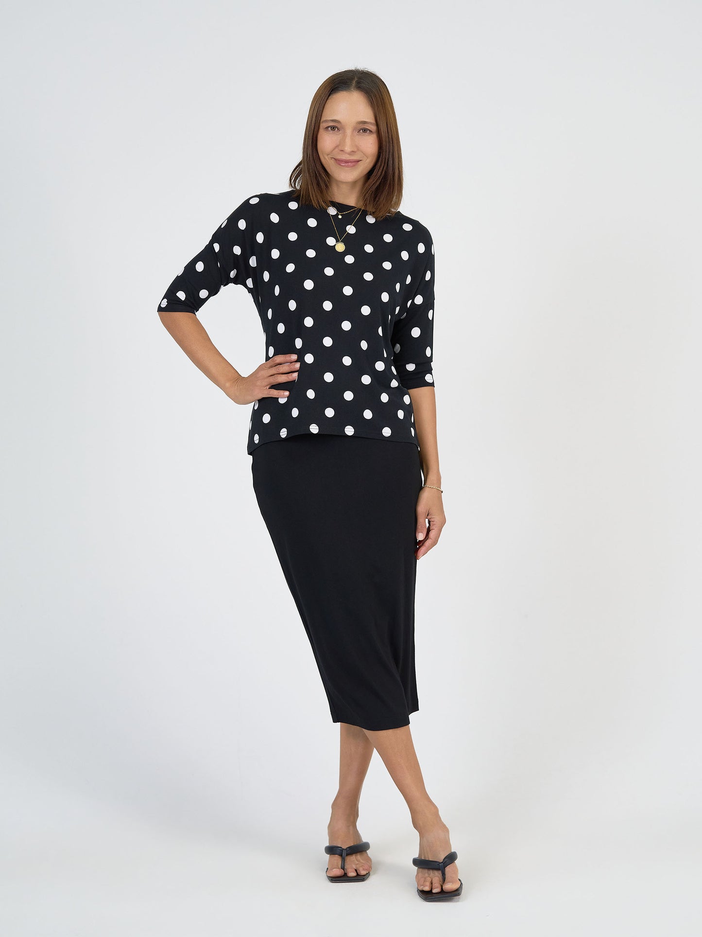 A woman stands against a white background wearing the Camila 3/4 Sleeve Bamboo Top - Polka Dot with a black midi skirt and black sandals. She has straight, shoulder-length hair and smiles with one hand on her hip.